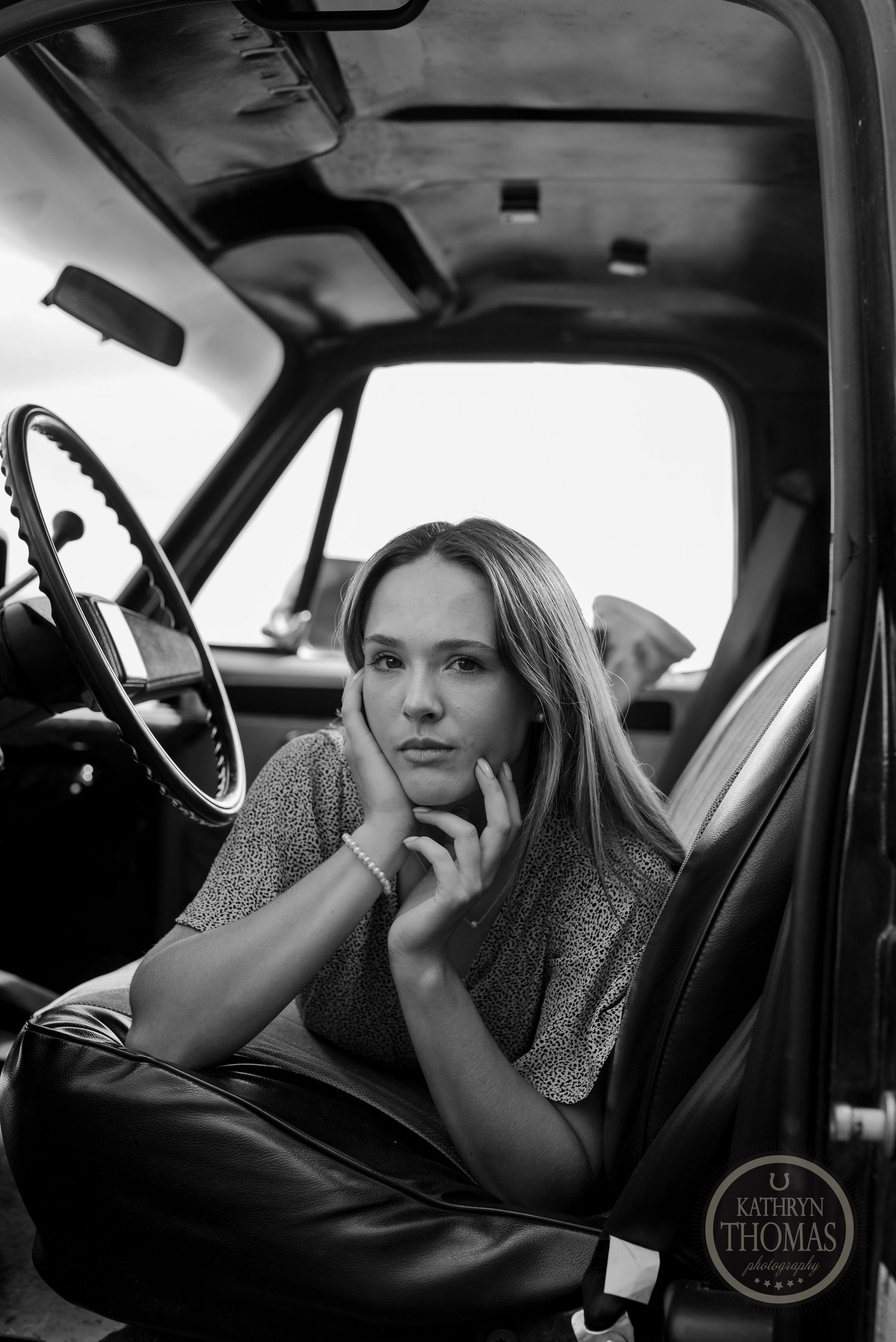 A moody black and white portrait of someone posing thoughtfully in a classic car interior.