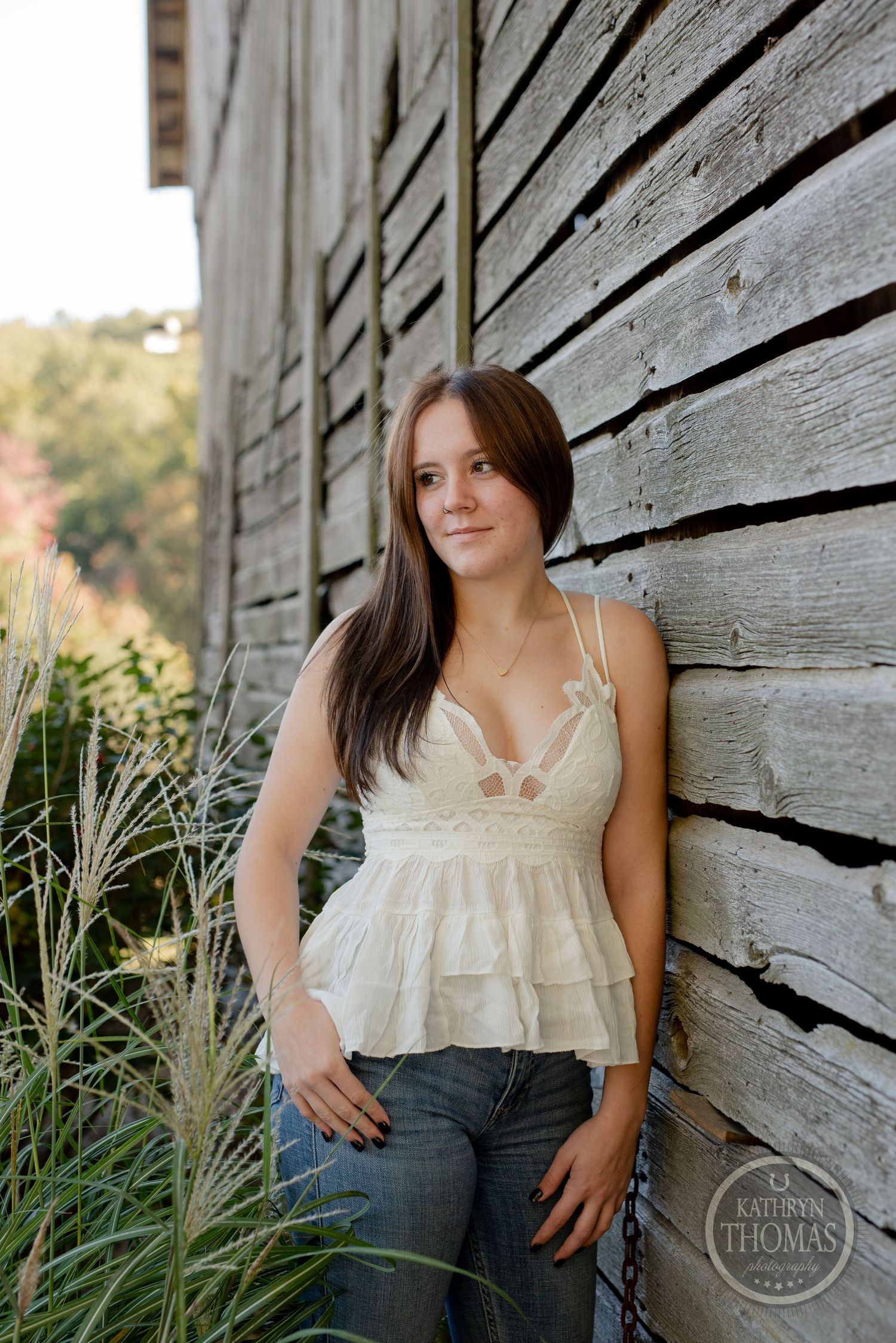 Portrait against rustic wooden wall wearing white tiered camisole top and jeans in outdoor setting.