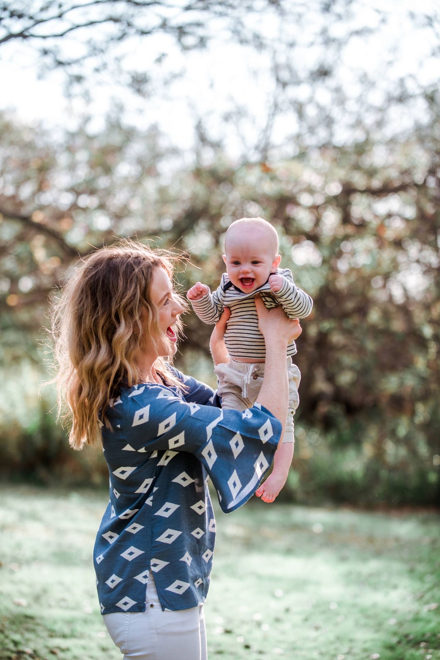 Parent in blue patterned dress lifts laughing baby wearing stripes in sunny outdoor setting.