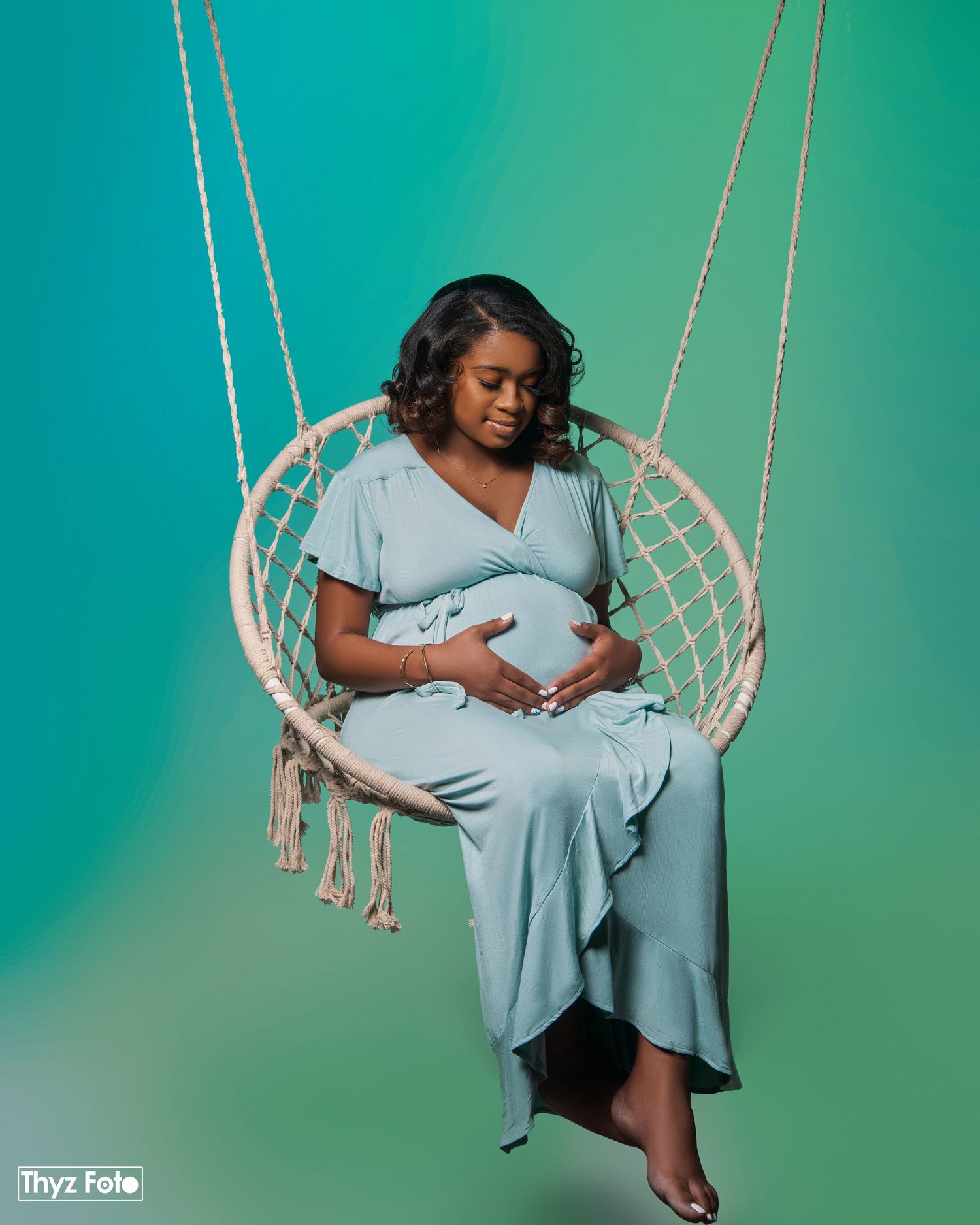 A pregnant woman in a pale blue dress sits peacefully in a hanging macrame chair against a teal backdrop.