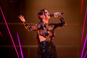 Concert performer in floral crop top and leather pants energetically singing on stage with dramatic lighting.