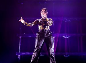 Performer in black leather pants and patterned top strikes powerful pose during concert.