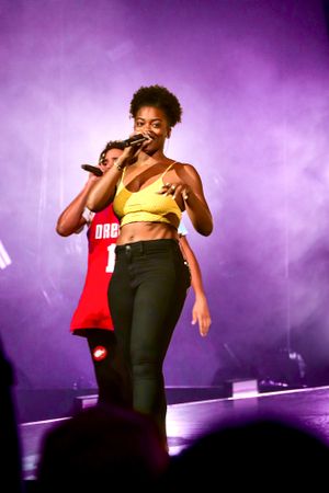 Performer in yellow crop top singing energetically on stage with purple lighting effects.