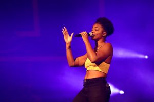 Performer in yellow crop top singing on stage with dramatic purple lighting.