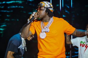 Performer in orange shirt wearing chains and bandana performs energetically on stage.