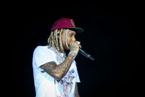 Performer in white t-shirt and burgundy cap performing with microphone against dark background.