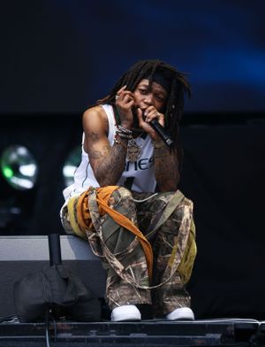 Performer in white tank top and camo pants crouches on stage during energetic performance.