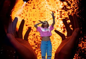 Performer in pink top and jeans against fiery orange backdrop with large silhouetted hands.