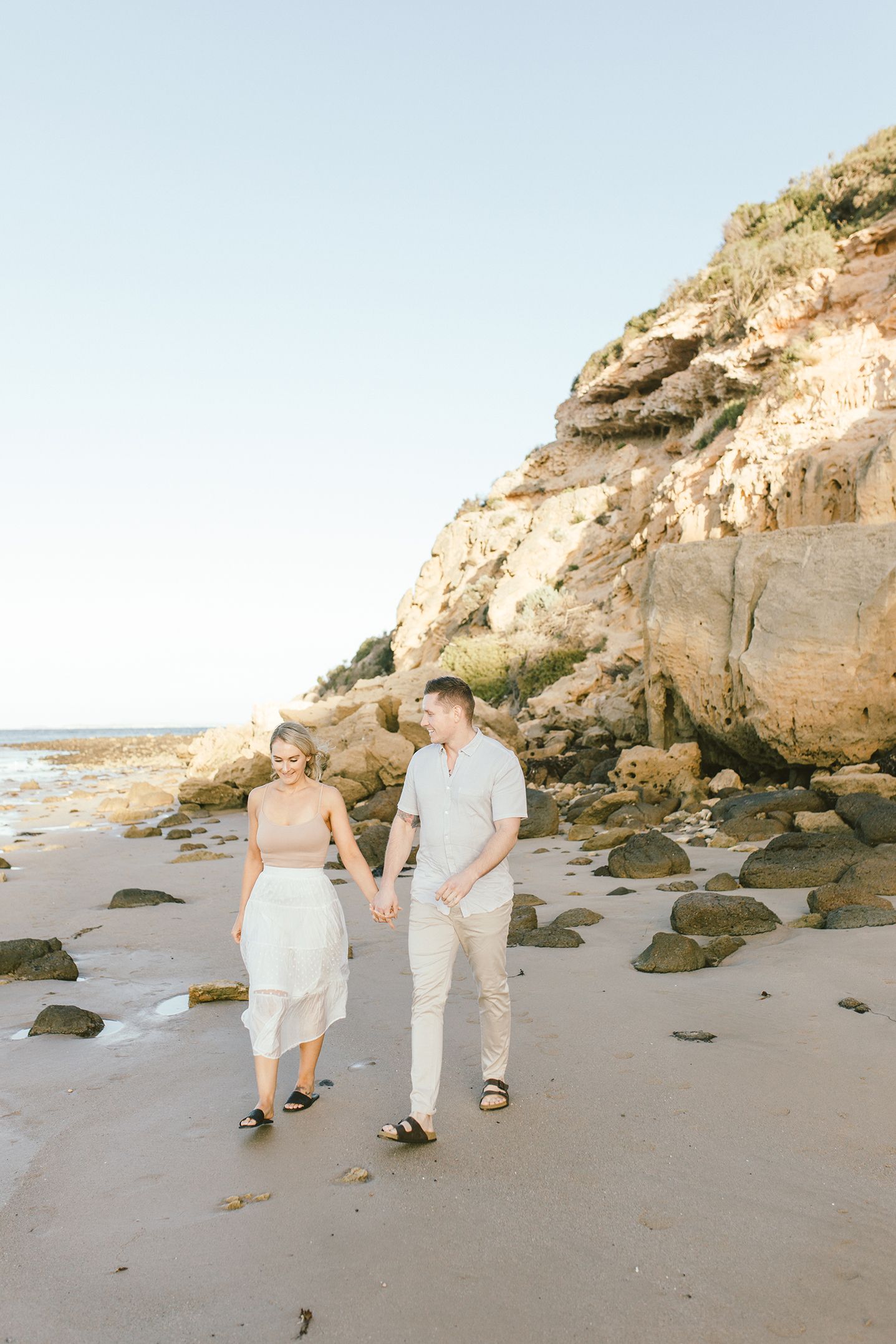 A couple takes a romantic stroll along a scenic beach with dramatic cliffs in the background.