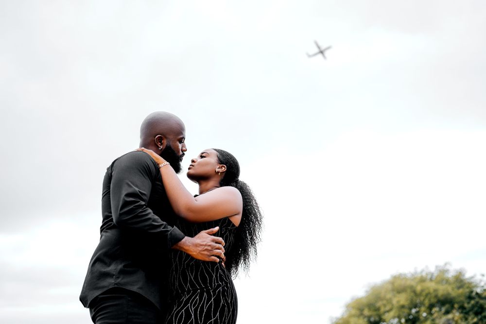 A couple shares an intimate moment outdoors with an airplane visible in the cloudy sky.
