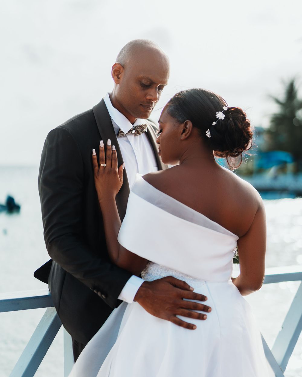 A romantic wedding couple shares an intimate moment on a beachfront balcony in formal attire.