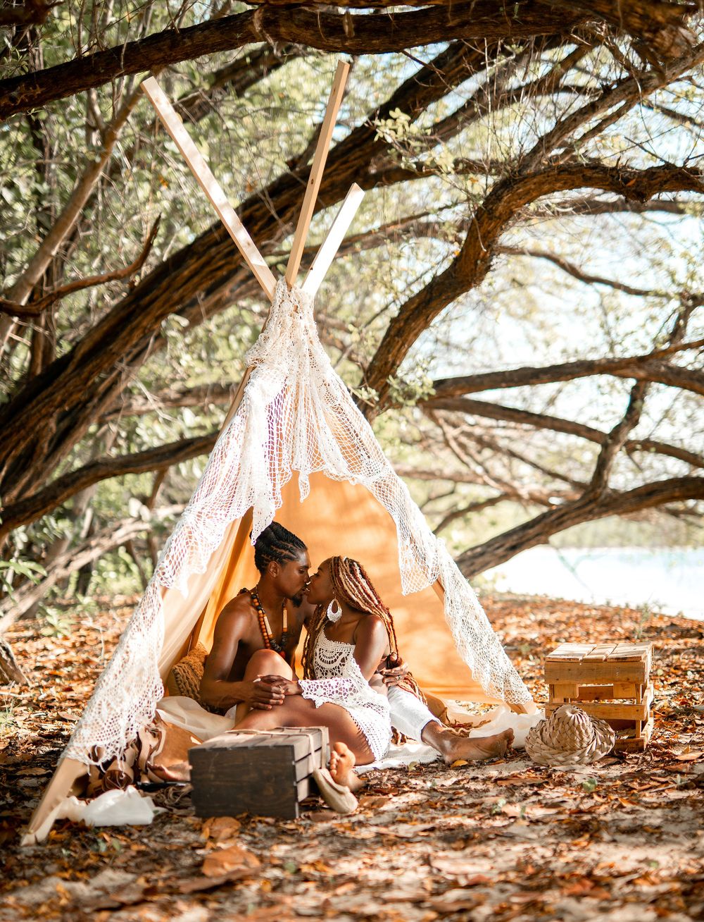 A romantic beach picnic setup with a macrame teepee under shady trees.