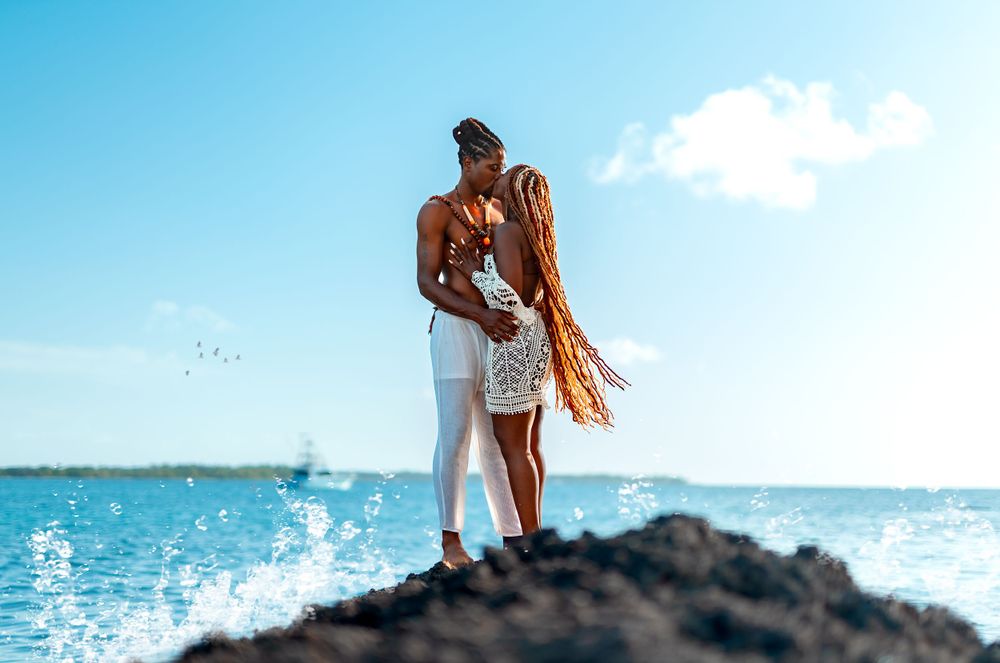 A couple embraces on rocky coastal outcrop with ocean waves crashing behind them.