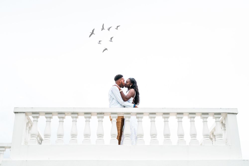 A romantic couple poses on a white balustrade with birds flying overhead.