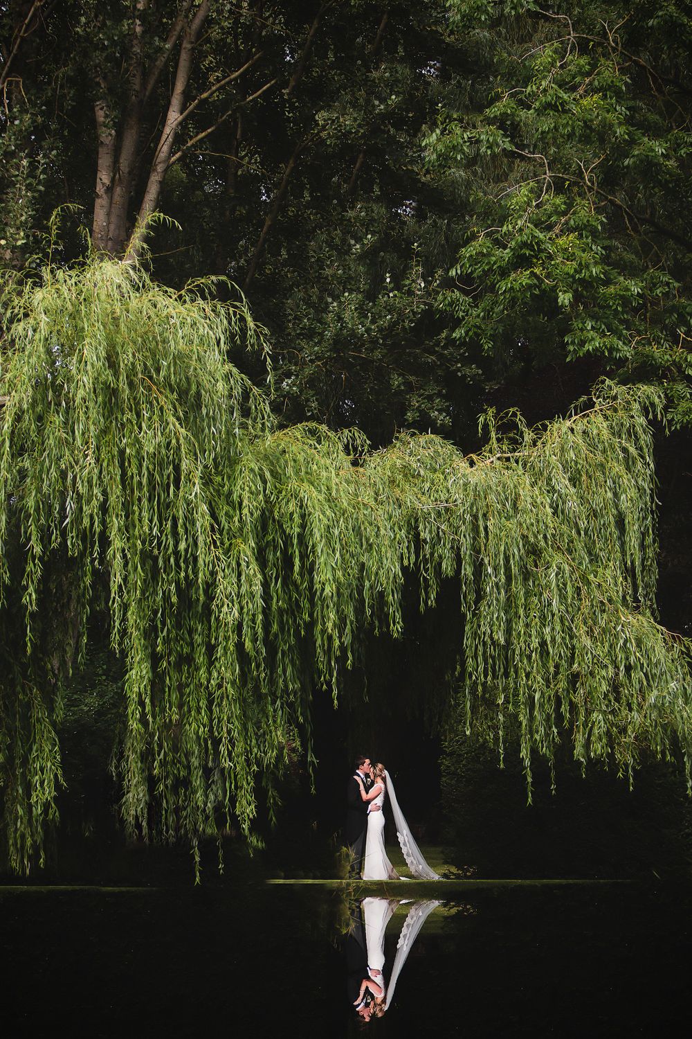 Romantic wedding couple stands beneath cascading willow trees reflected in still water at dusk.