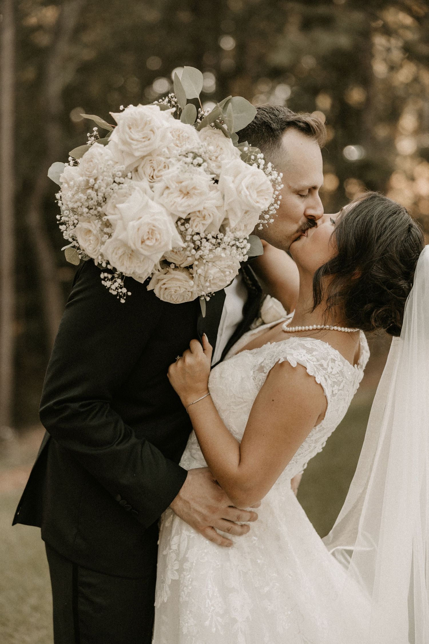 Romantic wedding kiss behind elegant white rose bouquet in forest setting.