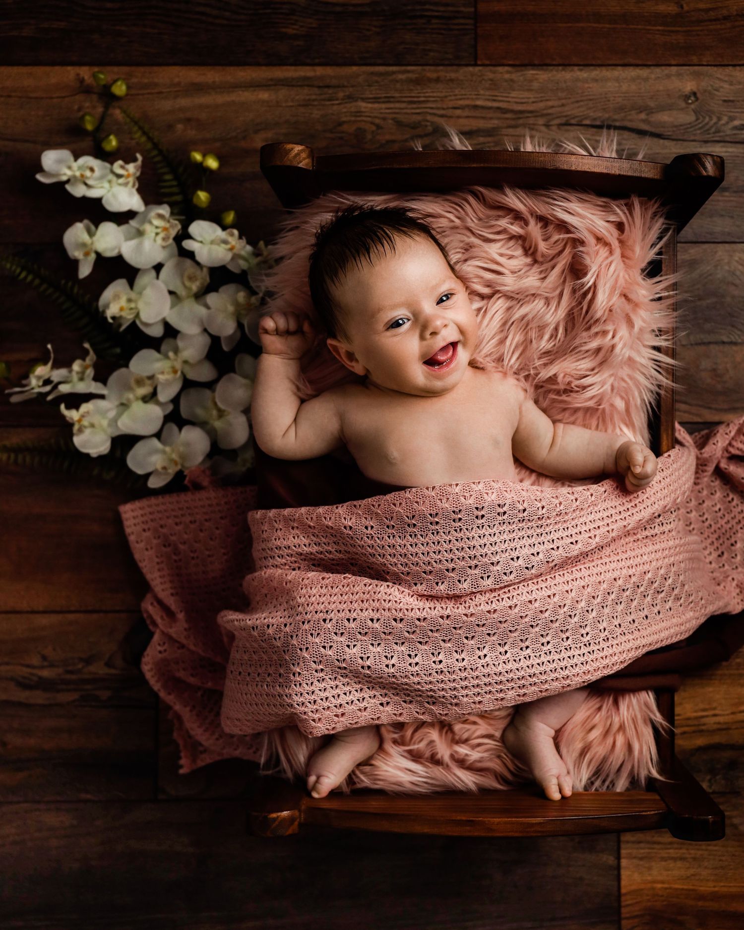 Baby girl smiling while lying on a small wooden bed with pink fur and white orchid flowers.