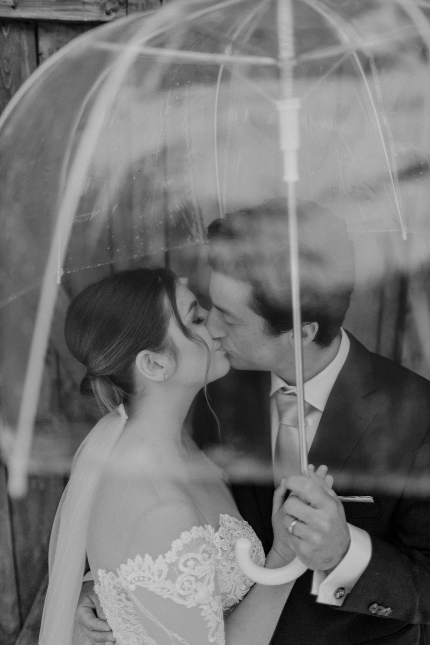 A couple shares a kiss under a clear umbrella in an artistic black and white portrait.