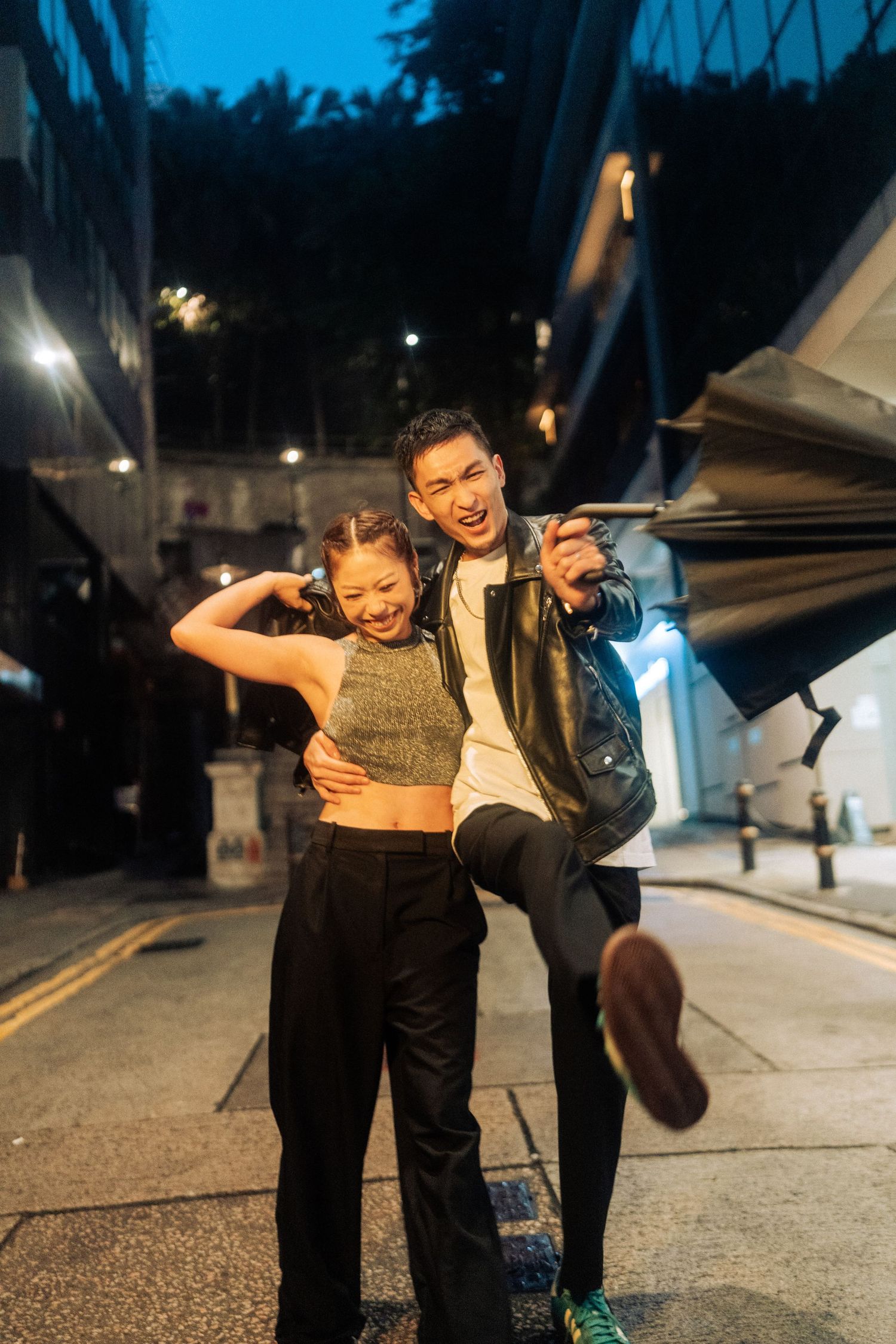 A candid nighttime photo of two people striking fun poses and laughing on an urban street.