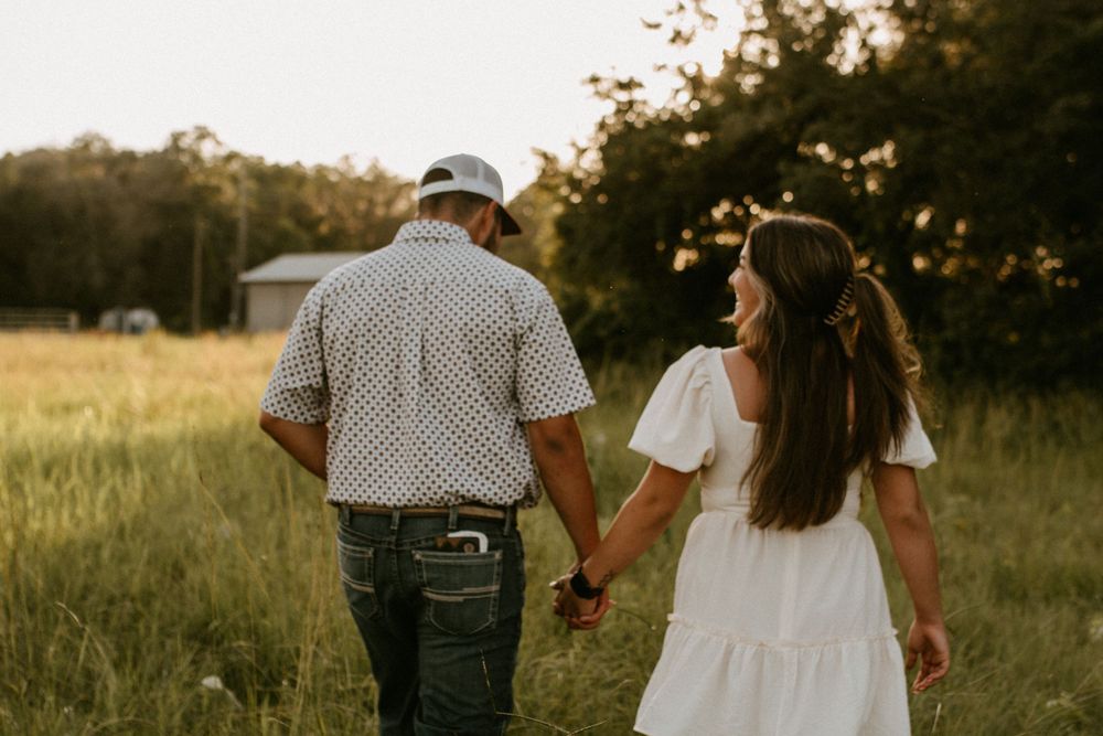 A couple walks hand in hand through a field at sunset.
