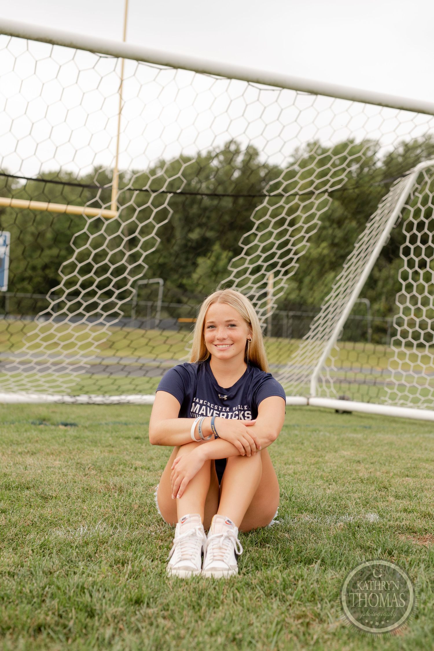 Soccer player sitting on grass field near goal post in navy uniform.