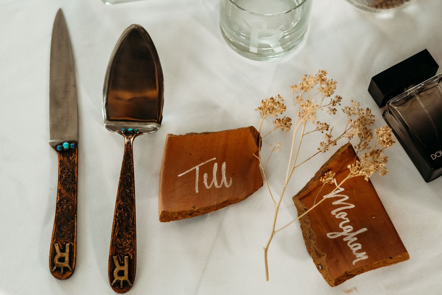 Elegant wedding styling details featuring dried flowers, brown leather accents, and handwritten calligraphy on a neutral surface.