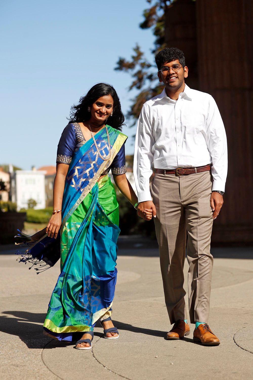 Couple in mixed traditional and modern attire walk hand in hand by classical architecture.