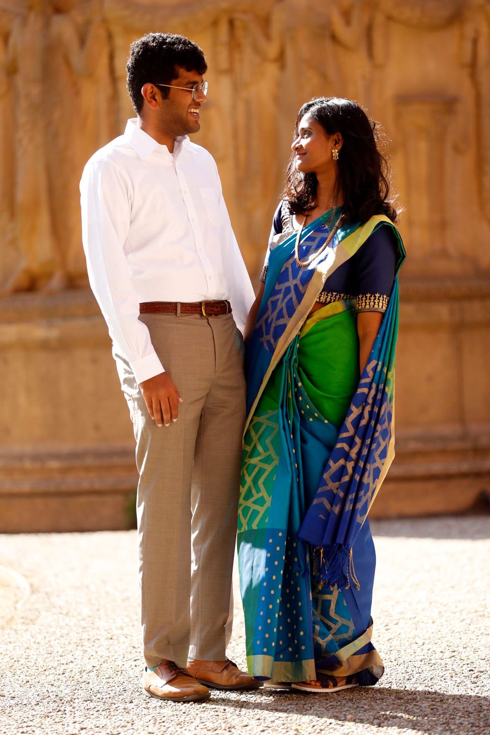 Couple in formal western and traditional Indian attire pose against ornate stone columns.
