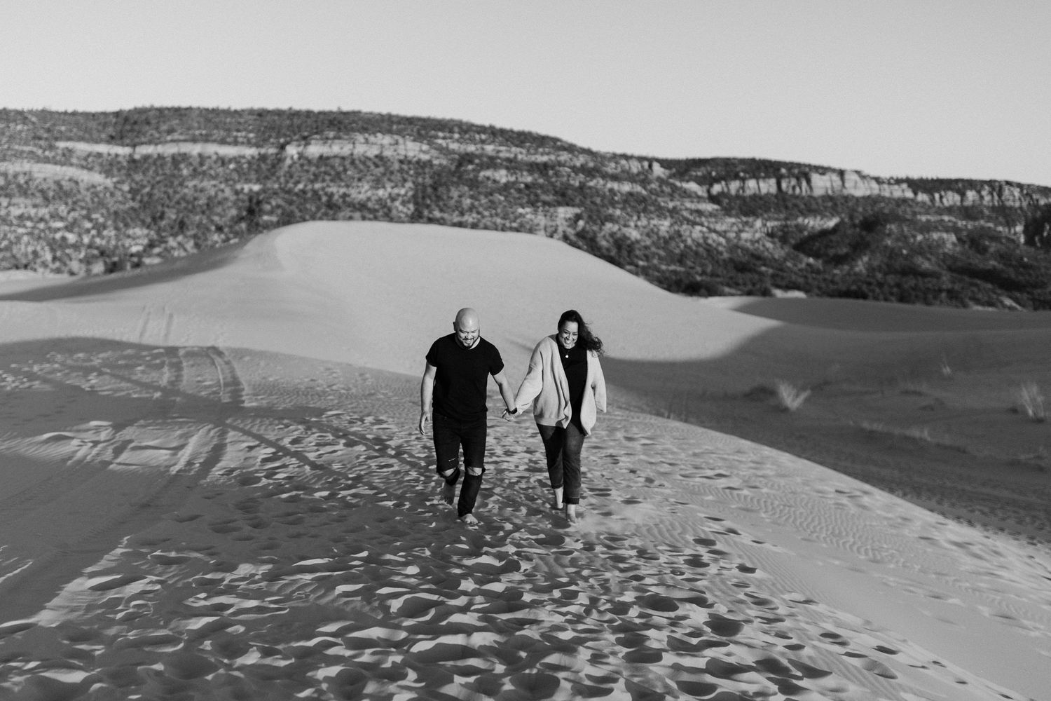 A couple explores vast sand dunes together against a mountainous backdrop in black and white.