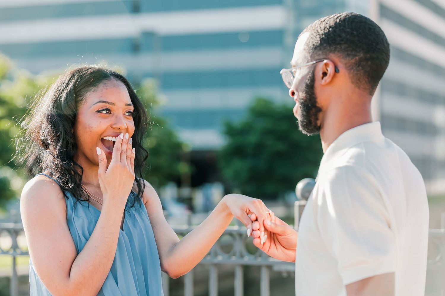 Man surprises woman with a marriage proposal outdoors as she covers her mouth in happy shock.