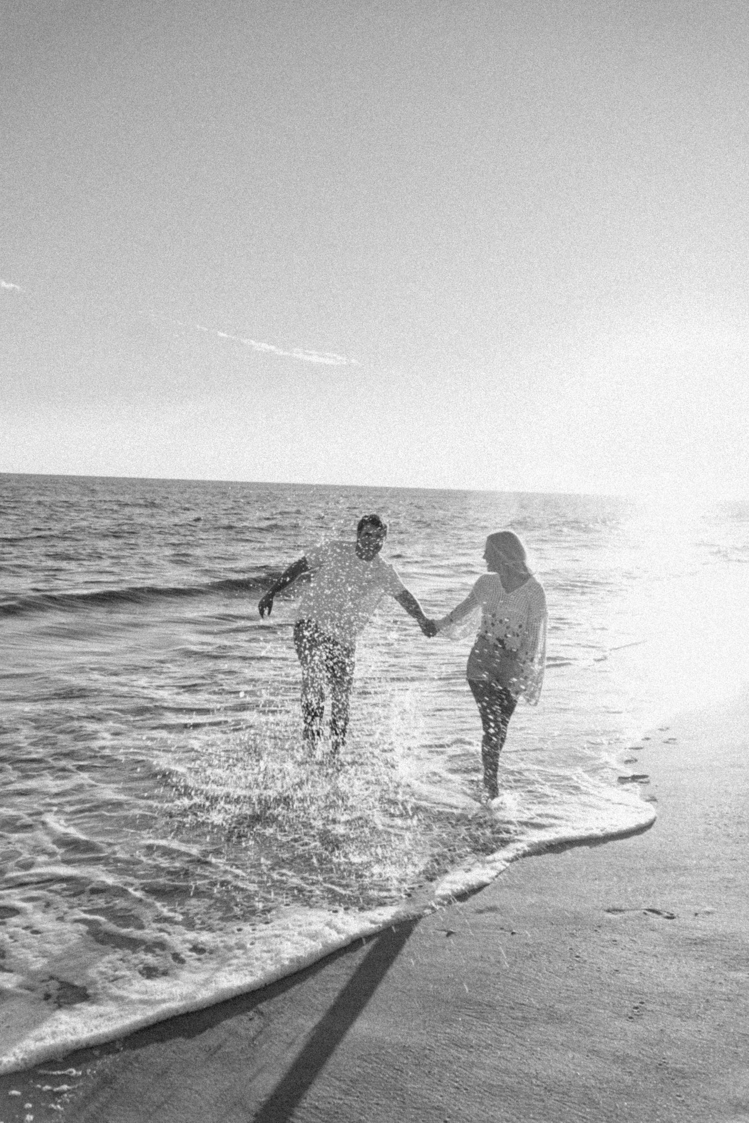 Black and white photo of couple splashing in ocean waves at sunset.