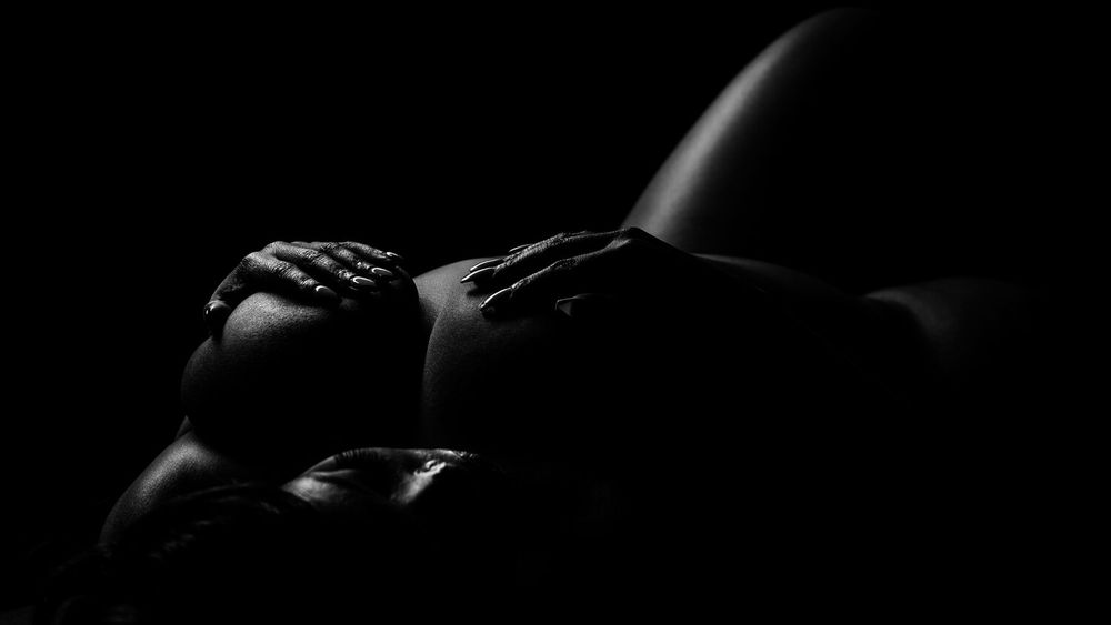Black and white artistic nude photograph with dramatic shadows highlighting curves and contrasts against dark background.
