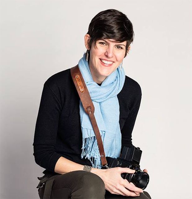 Professional photographer wearing a blue scarf and black sweater holds camera with strap.