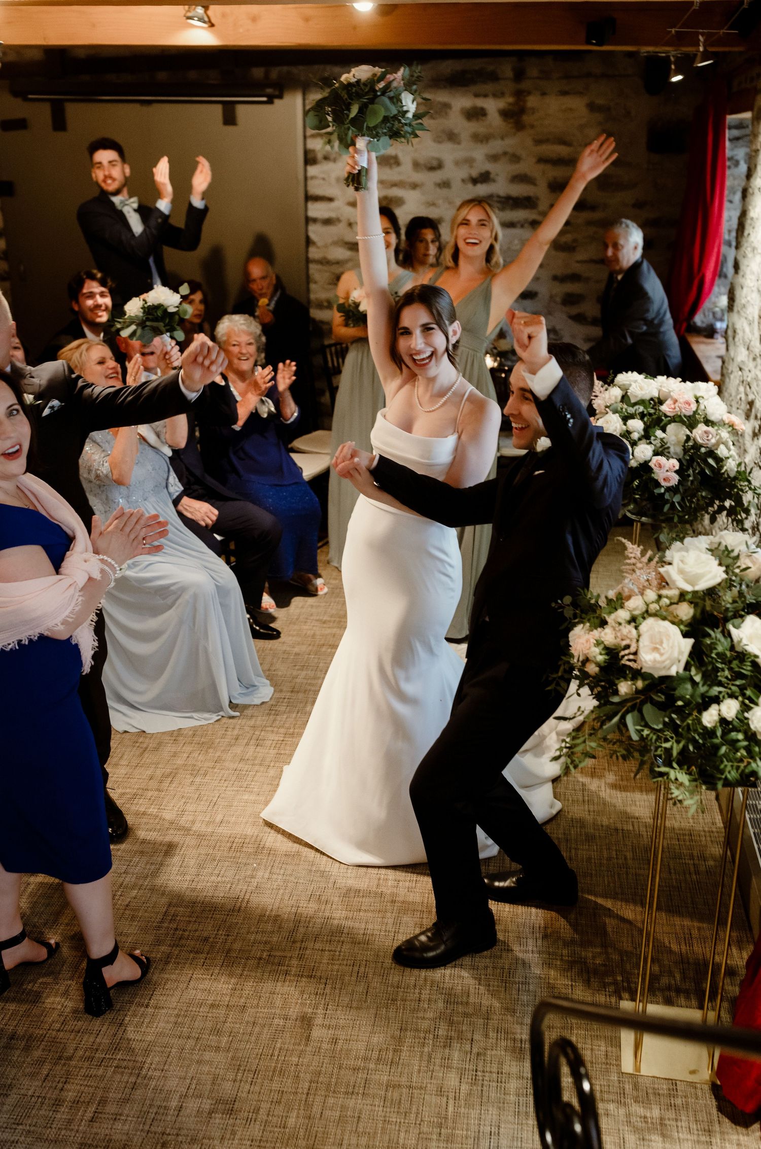Couple celebrates with energetic dance moves during wedding reception in rustic venue.