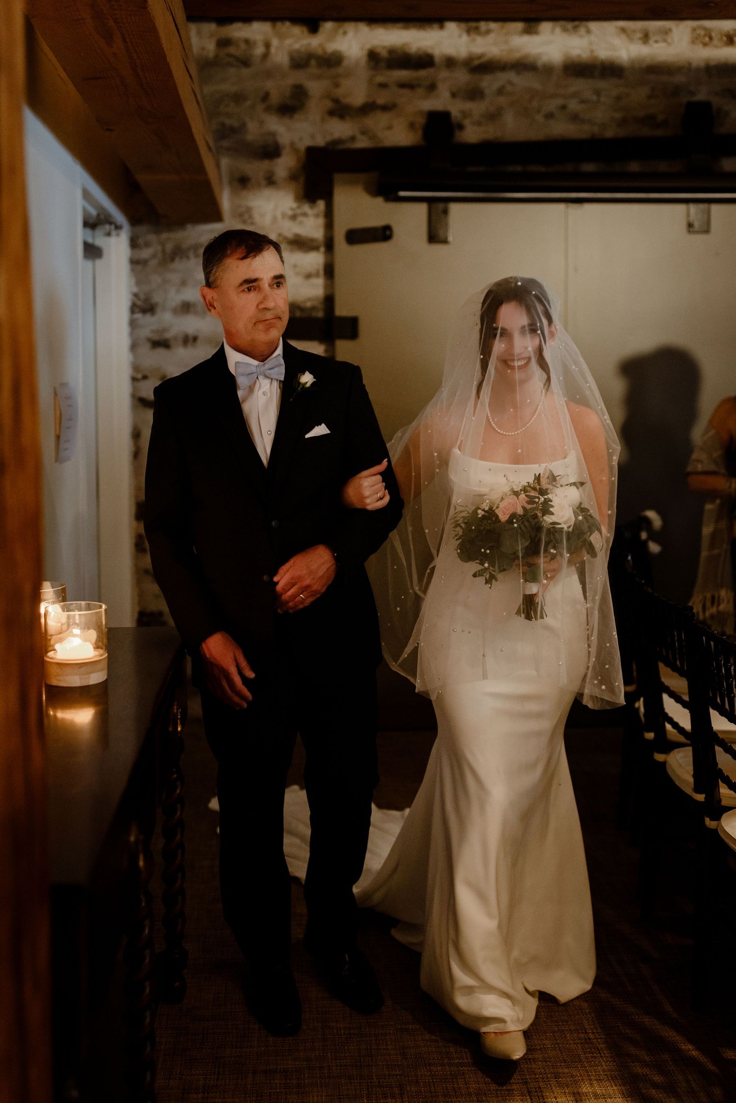 Bride in elegant white gown walks down candlelit aisle in rustic venue.