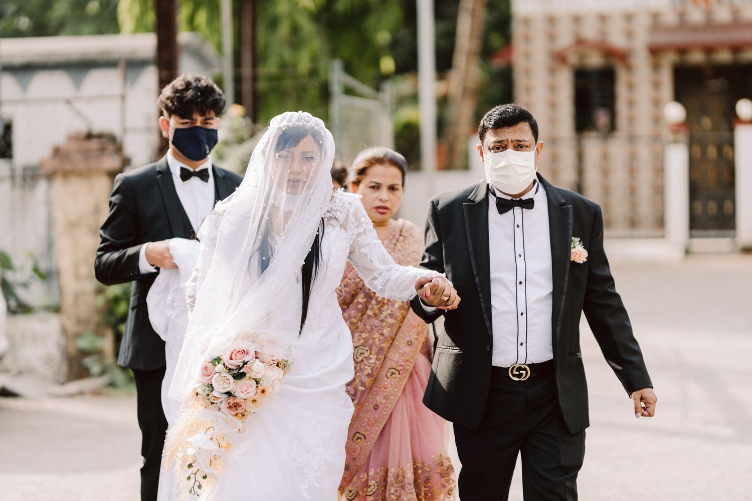 A wedding ceremony procession walking together outdoors with guests wearing formal attire and face masks.