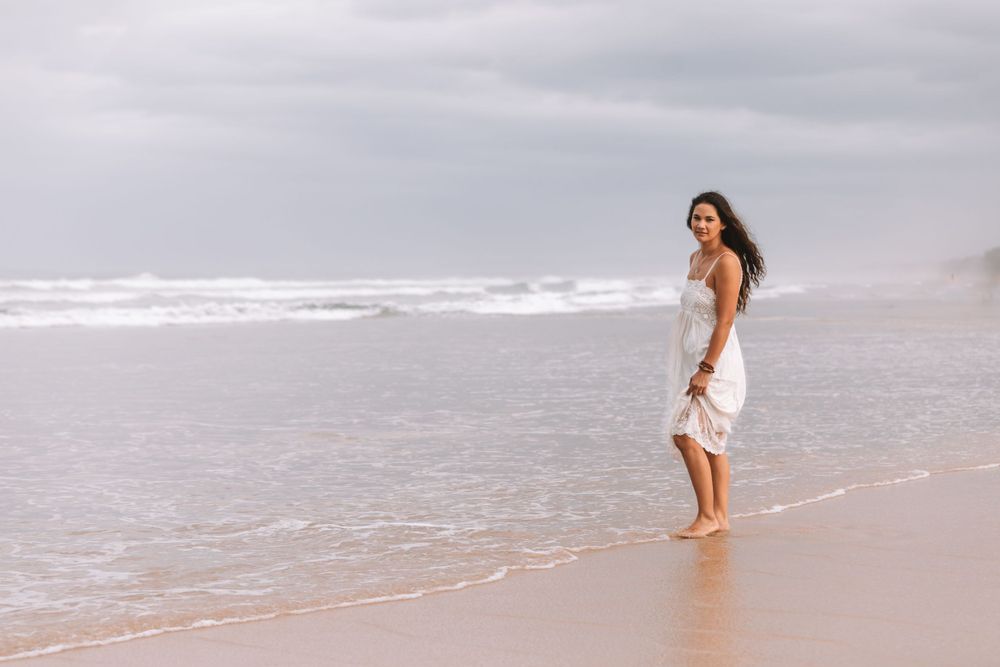 Woman in white dress walking barefoot along a misty beach shoreline as waves roll in on an overcast day.
