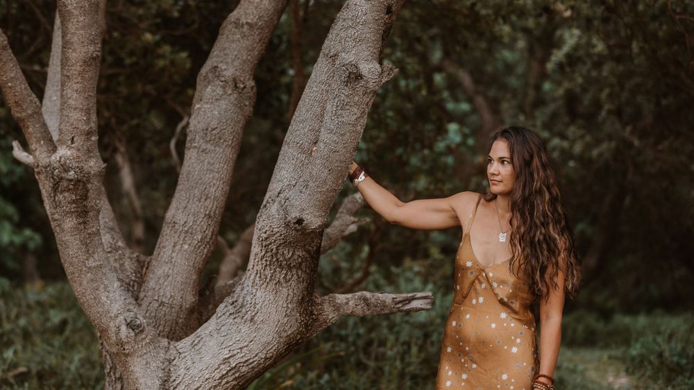 Woman in golden dress poses near a large branching tree trunk in a dreamy outdoor forest setting.