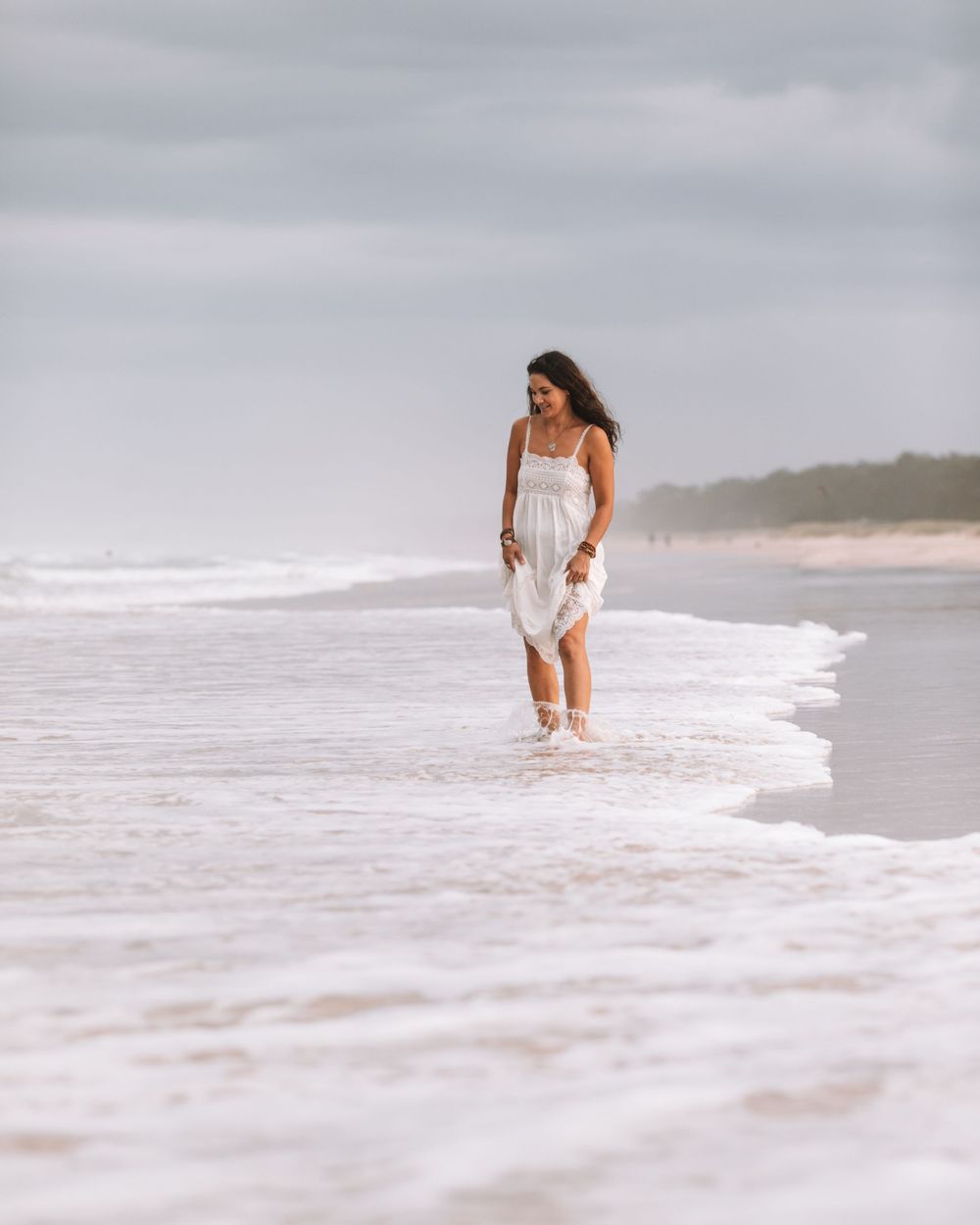 Person in a white dress walking along a misty beach shoreline as waves roll in on an overcast day.