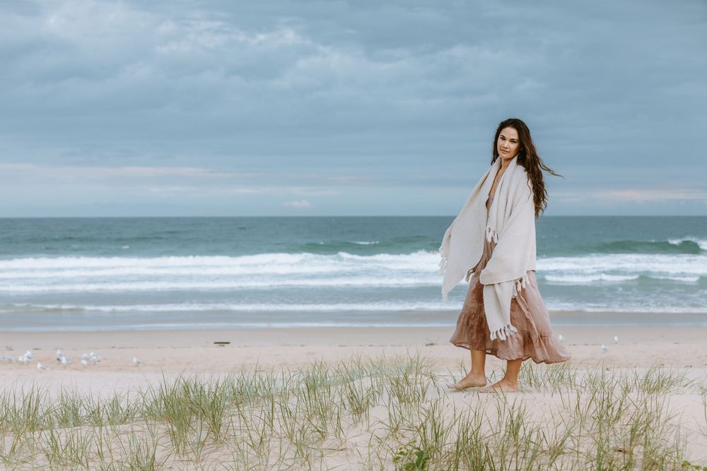 A person in a flowing white dress walks along a sandy beach with ocean waves and cloudy skies in the background.