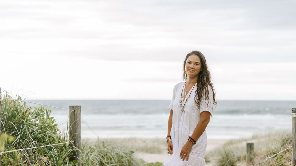 Person in flowing white dress stands near weathered wooden fence post along coastal beach path on cloudy day.
