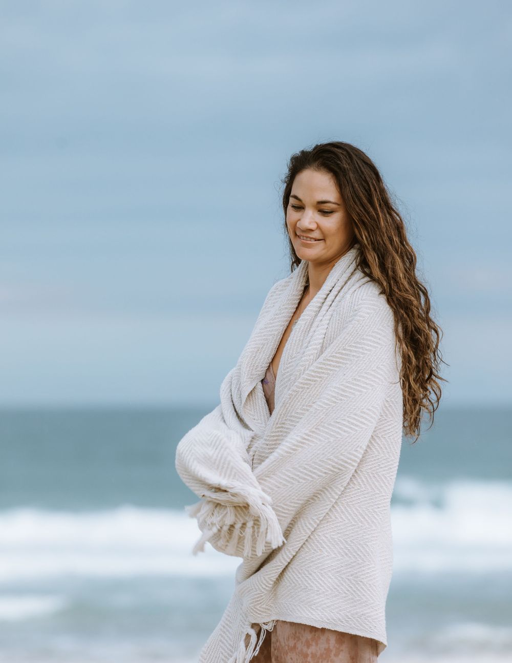 Person wearing a cozy white cardigan stands peacefully near ocean waves on a cloudy day at the beach.