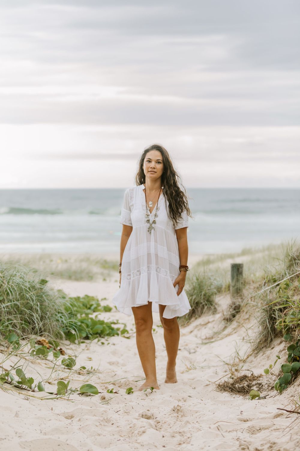 A person in a flowing white summer dress walks barefoot along sandy beach dunes on an overcast day.