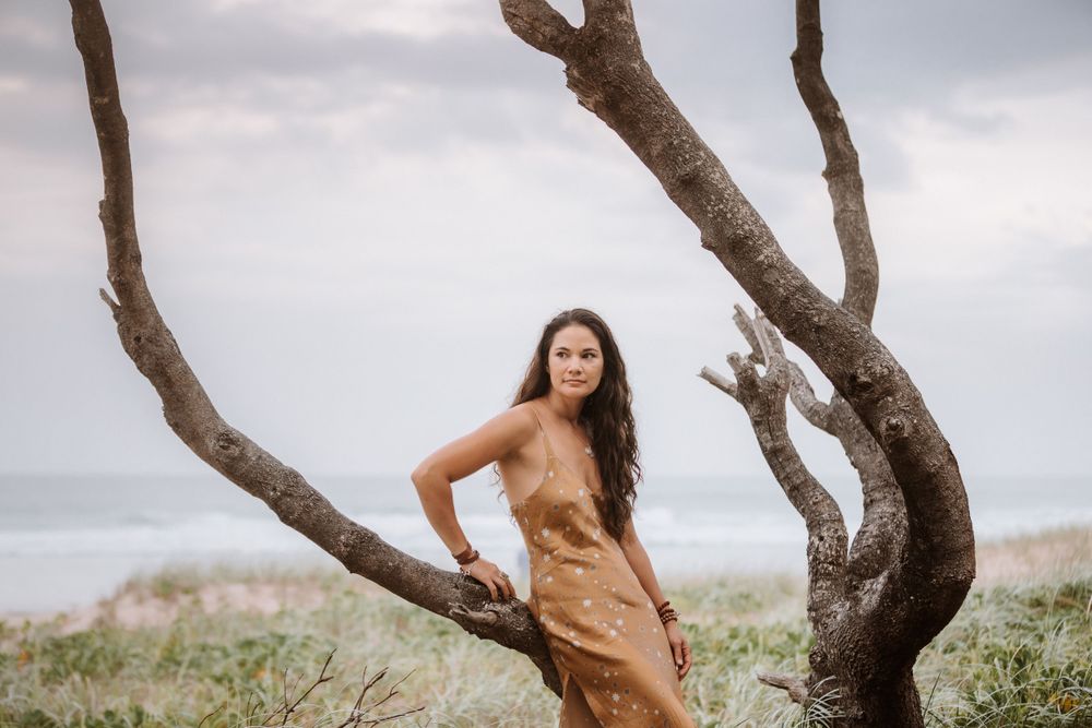 A person in a brown dress poses against a twisted driftwood tree trunk on a misty coastal beach.