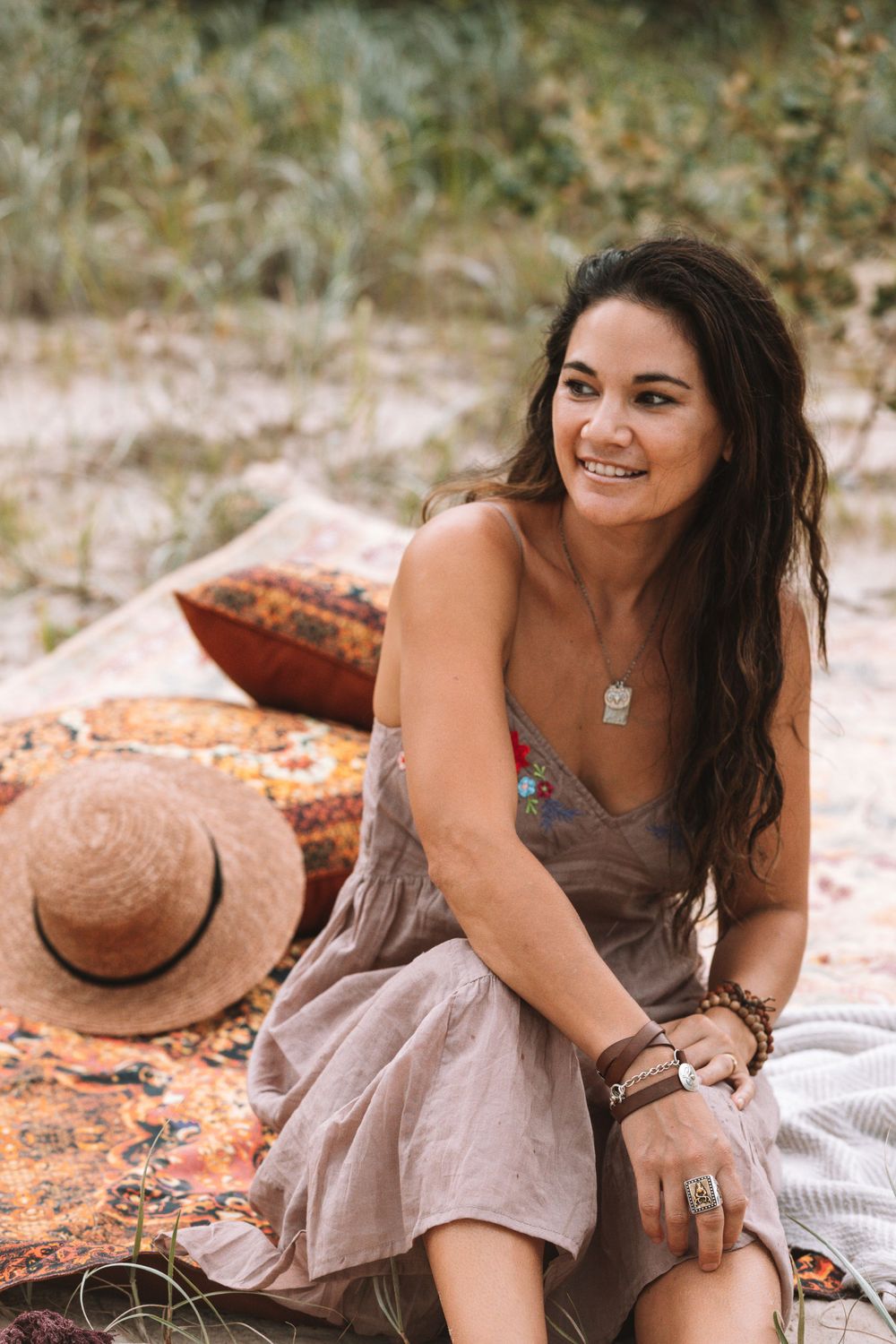 A woman in a beige dress sits on a bohemian blanket with a straw hat nearby during a sunny outdoor photoshoot.