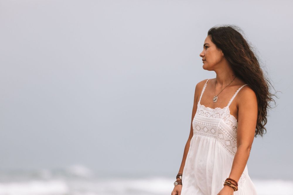 Person with long dark hair wearing a white sundress stands on a misty beach while looking to the side.