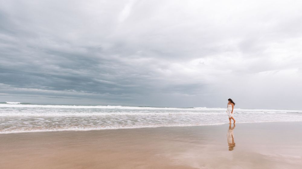 A solitary figure walks along a serene sandy beach under moody clouded skies with crashing waves in the distance.