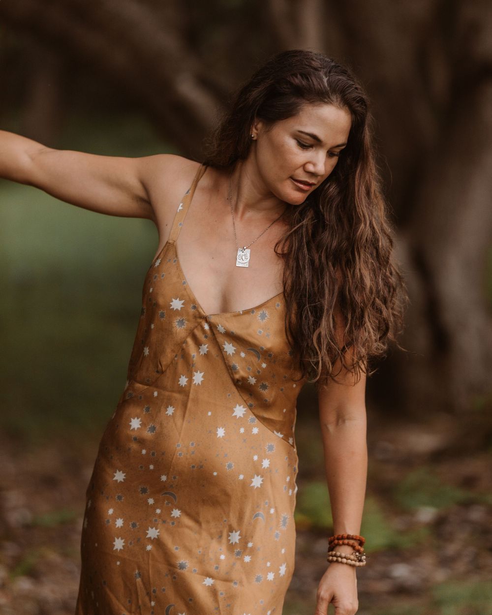A woman in a warm brown polka dot sundress poses outdoors with flowing long hair and delicate jewelry.