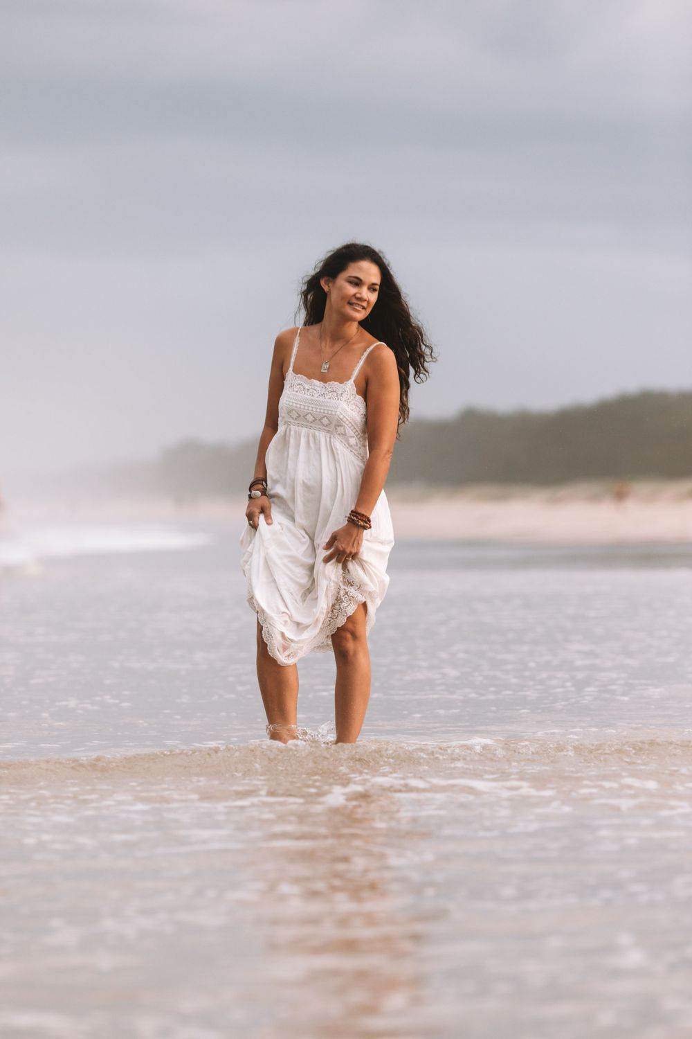 Woman in flowing white dress enjoying the beach as waves splash around her feet on an overcast day.