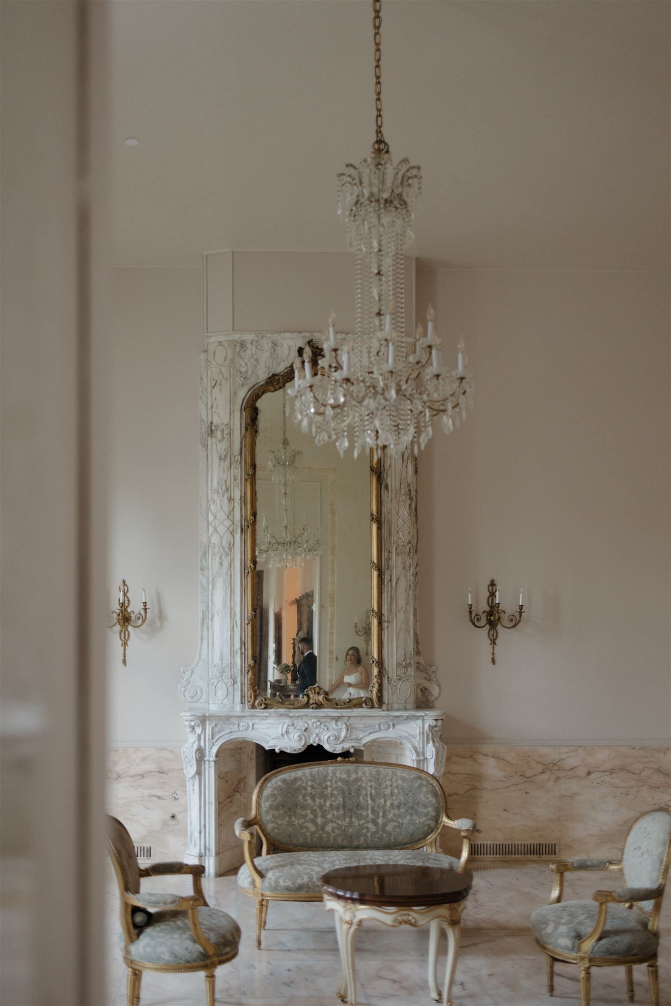 Elegant French-style room with ornate crystal chandelier, antique mirror on marble mantle, and vintage upholstered chairs.